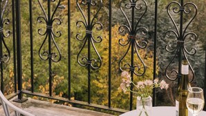 A balcony with wrought iron railings, a table with a bottle of wine and glasses, and a vase with flowers.