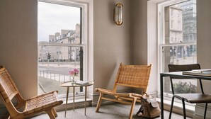 A room with two wicker chairs, a small round table with a vase of flowers, and a view of the city through large windows.