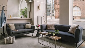 A modern living room with two dark sofas, a small coffee table, and a side table. There is a coat rack with a jacket and a handbag, and a potted plant on the windowsill.