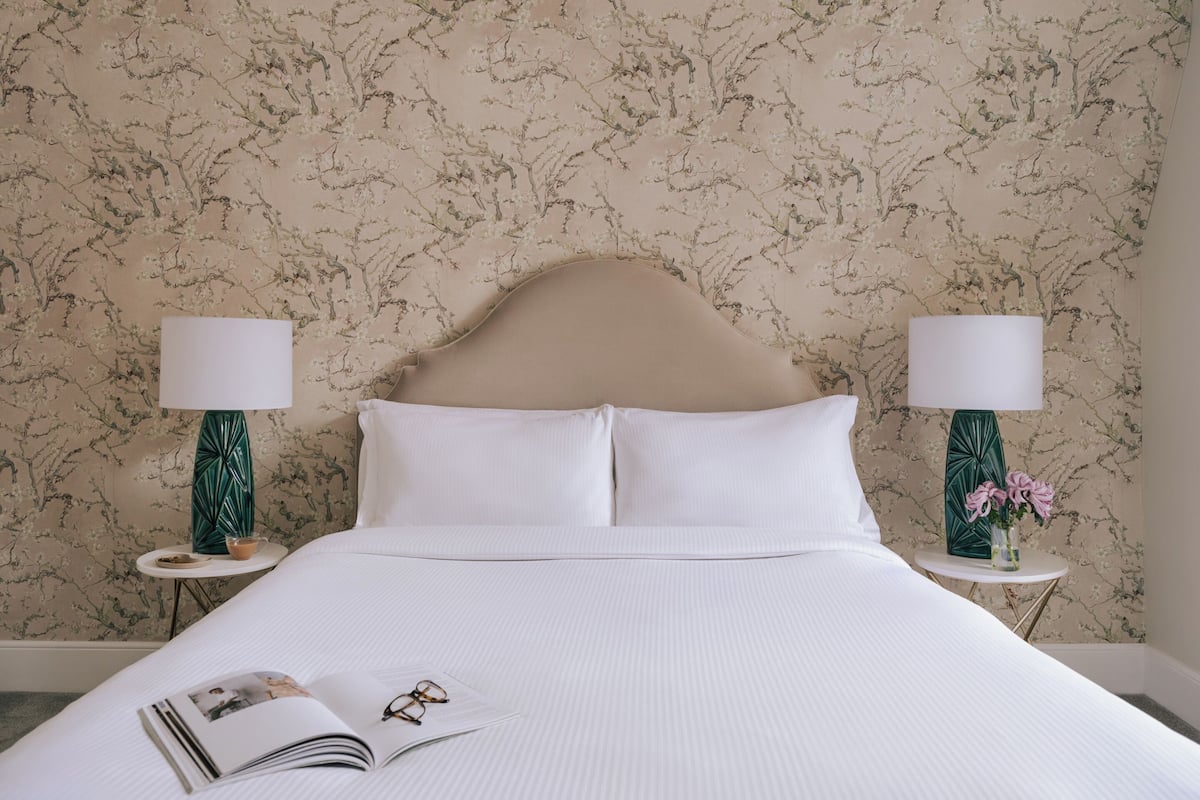Una cama bien hecha con sábanas blancas, dos mesitas de noche con lámparas y un estampado floral en el papel tapiz.