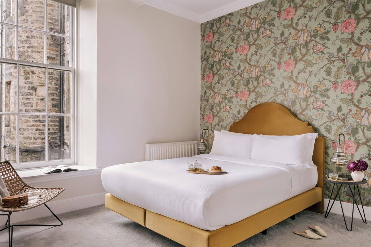 Una cama bien hecha con cabecera, una ventana con vista a un edificio de piedra, una silla con asiento de mimbre y una mesita con lámpara y un jarrón de flores.