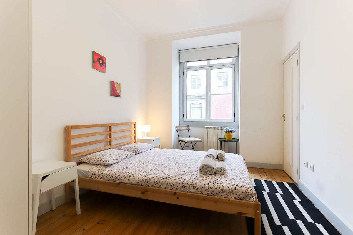 Quarto com cama de madeira, mesa de cabeceira pequena, cadeira, janela com vista para edifícios e porta.