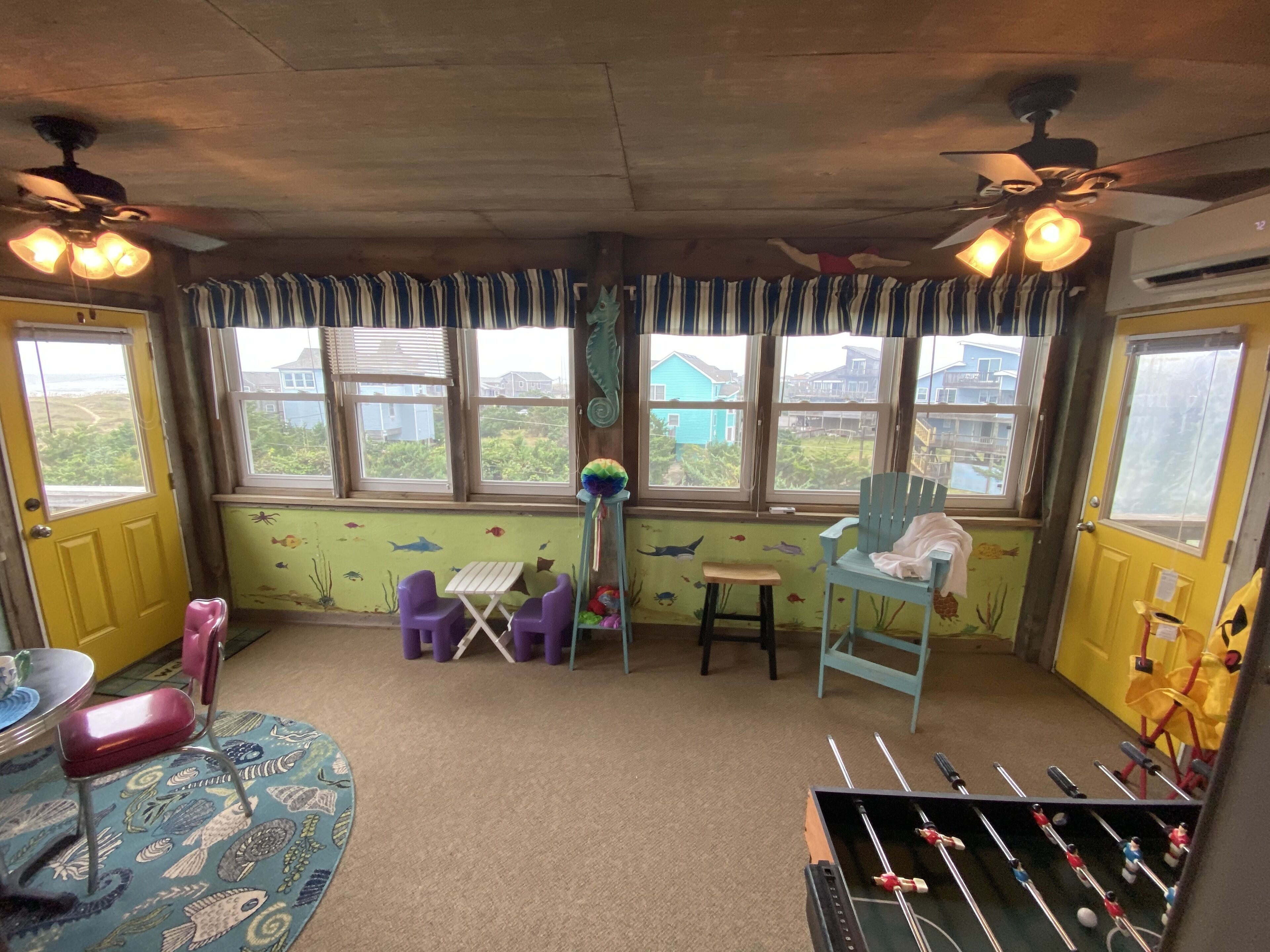 A room with a window view, a small table, two chairs, a foosball table, and a ceiling fan.