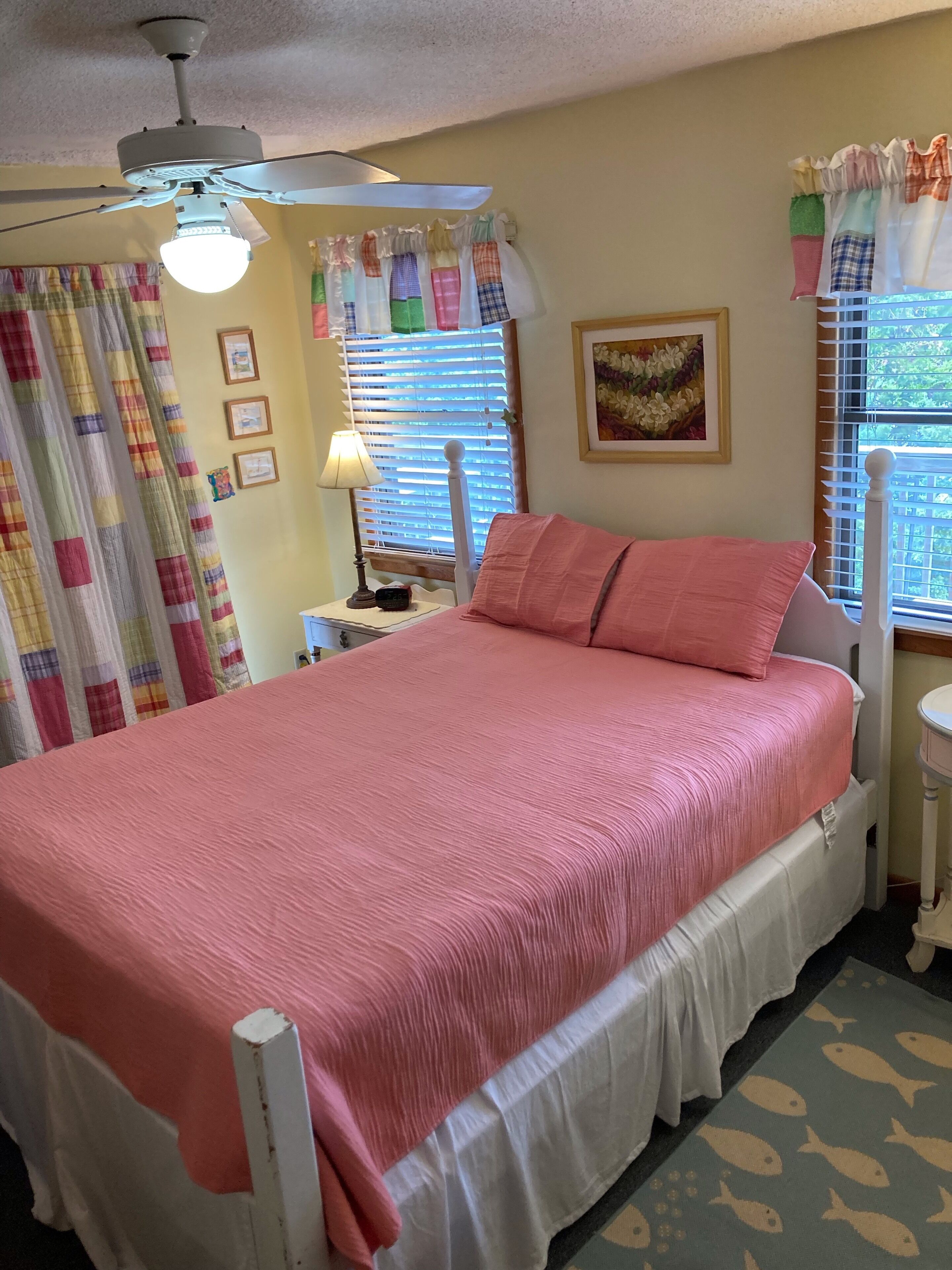 A bedroom with a bed, a ceiling fan, a window with blinds, and a wall with framed pictures.