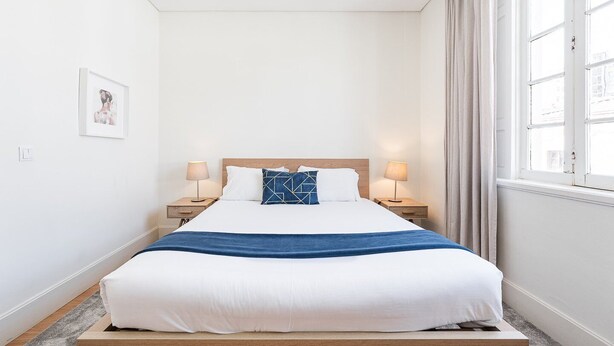 Un lit bien fait, avec une literie blanche et bleue, deux tables de chevet avec des lampes, un tableau encadré au mur et une fenêtre avec des rideaux.