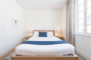 Een netjes opgemaakt bed met wit en blauw beddengoed, twee nachtkastjes met lampen, een ingelijst foto aan de muur, en een raam met gordijnen.