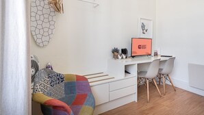 Une chambre moderne et minimaliste, avec un bureau blanc, un écran d’ordinateur affichant le message « PRÊT POUR L’ARRIVÉE DES CLIENTS », une chaise, un canapé coloré à motifs matelassés et un porte-serviettes fixé au mur.