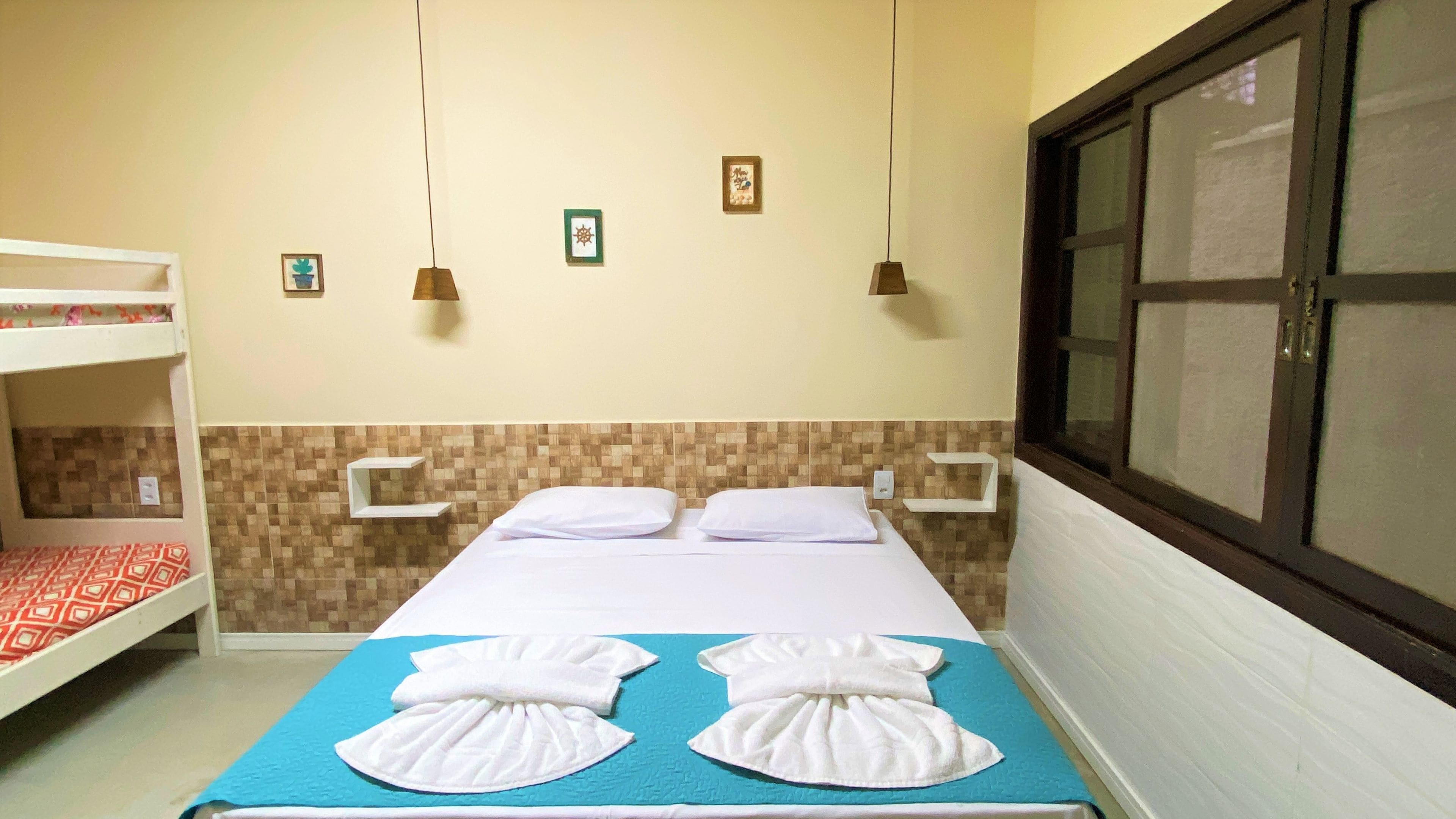 A hotel room with a bunk bed, a single bed with towels, a window with blinds, and wall-mounted lights.
