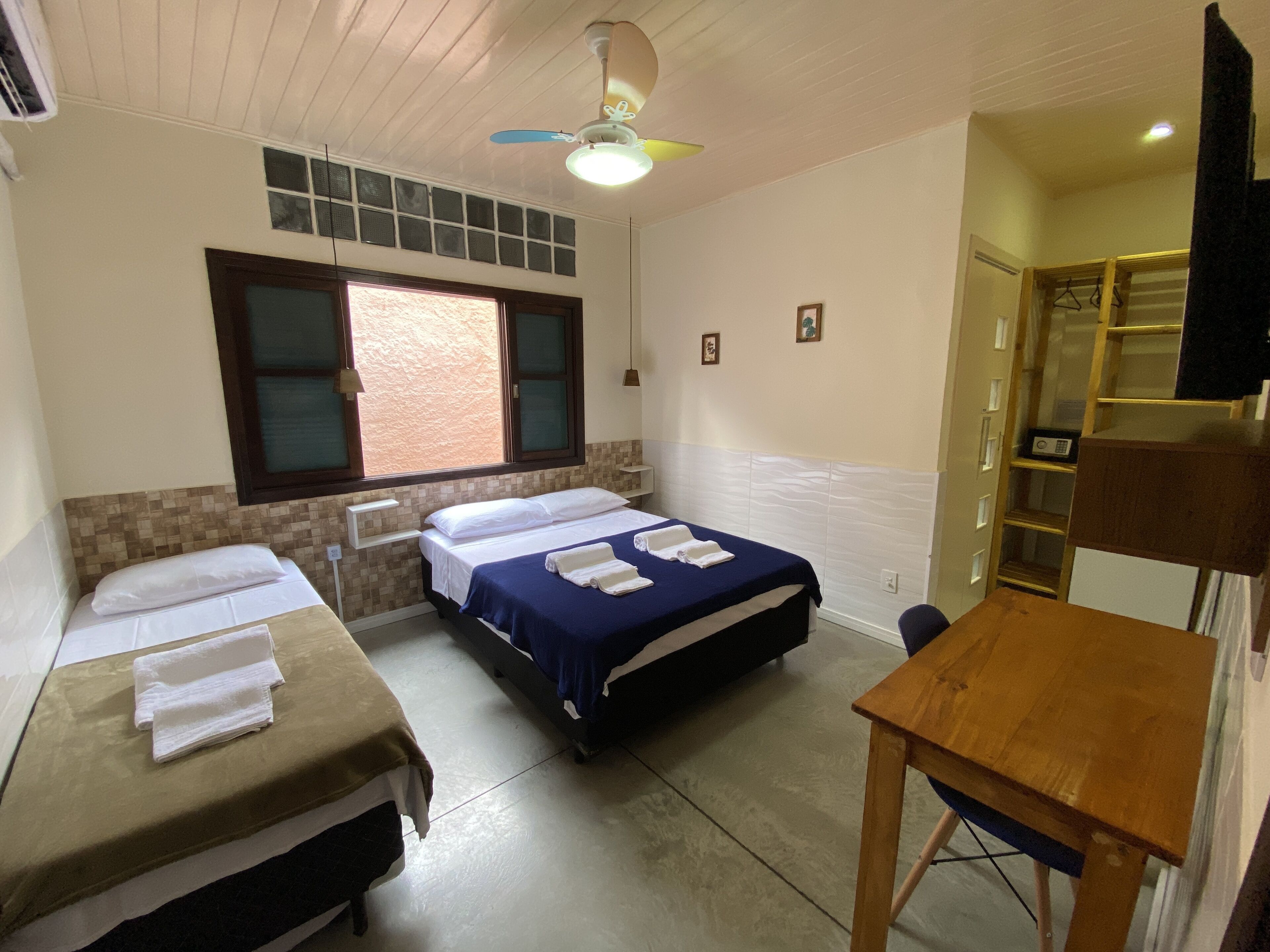 A room with two beds, a wooden table, a ceiling fan, and a window with blinds.