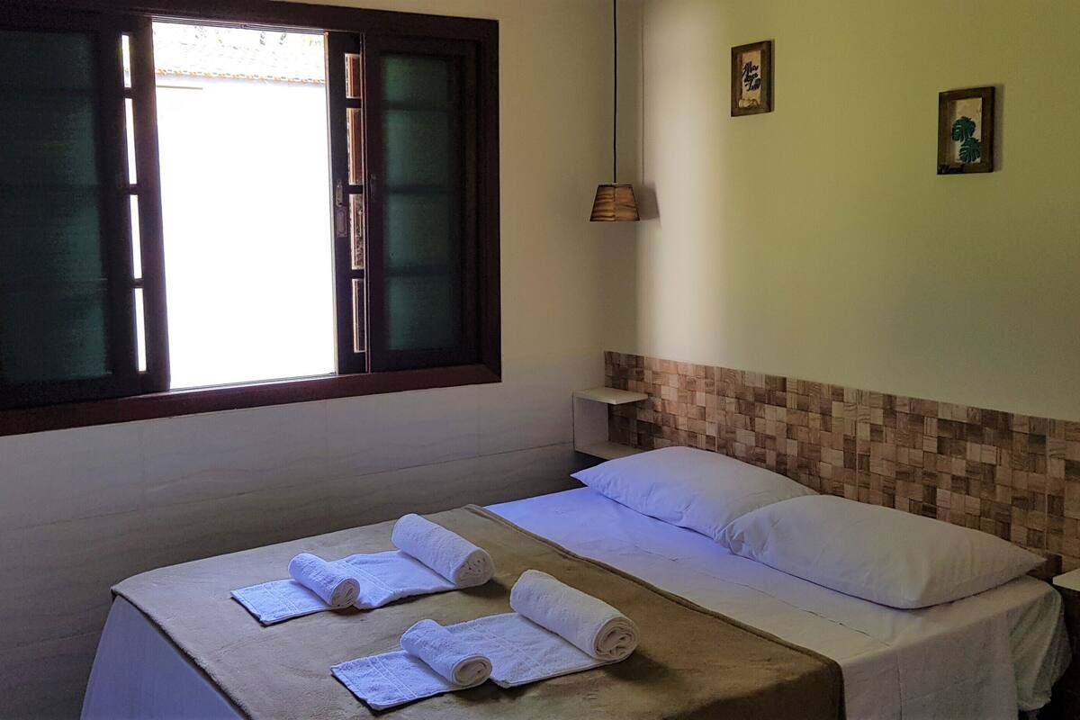 Un dormitorio con cama, dos almohadas, una ventana con contraventanas de madera, una mesita de noche y un estante montado en la pared.