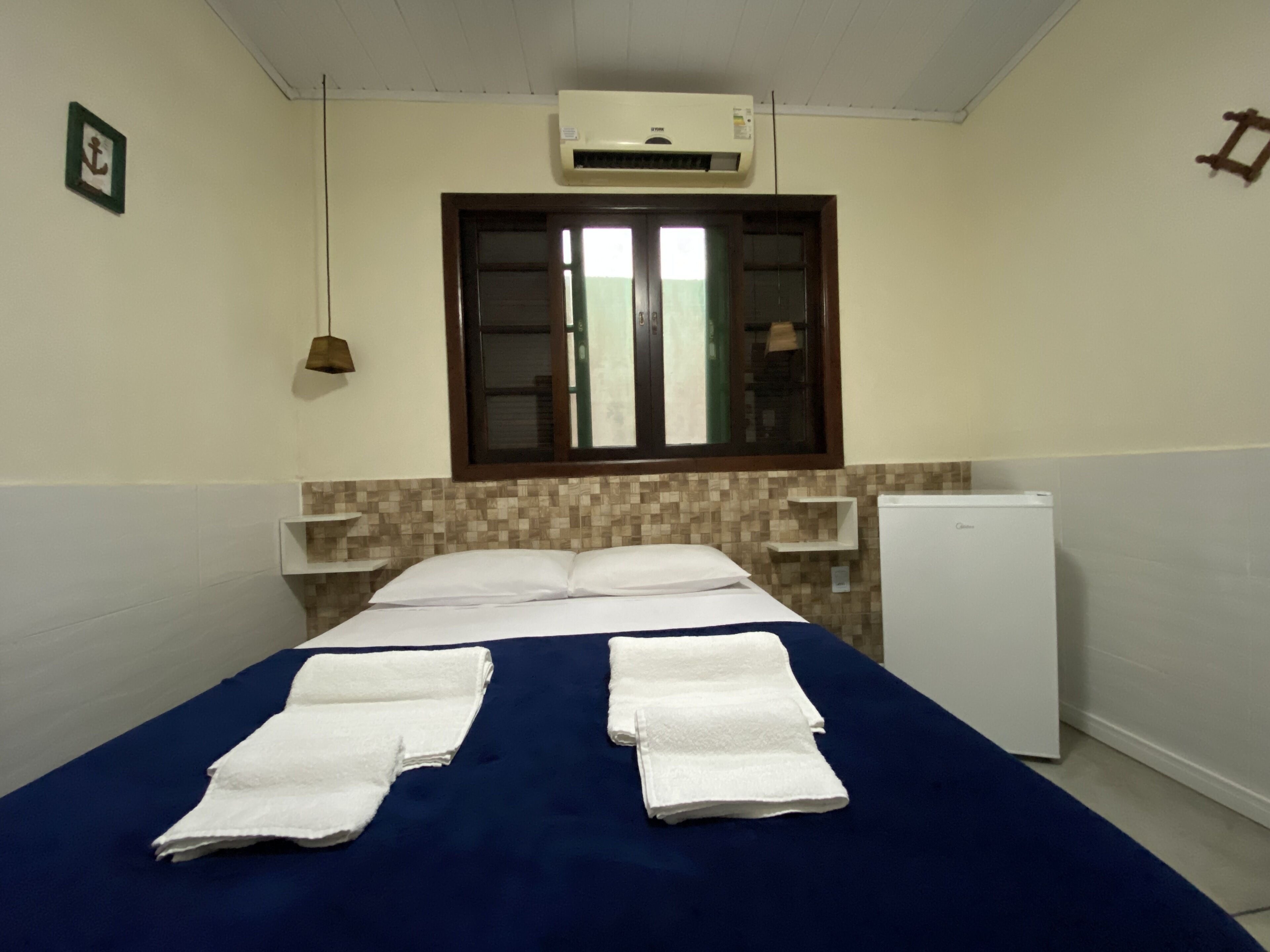 A hotel room with a bed, a window, a wall-mounted air conditioner, a small refrigerator, and a towel rack.
