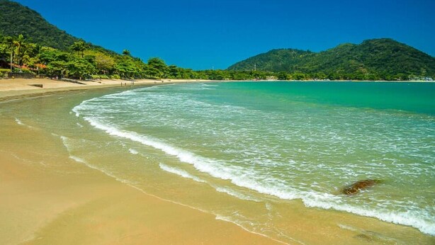 Una playa con agua turquesa cristalina, orilla arenosa y colinas verdes exuberantes al fondo.