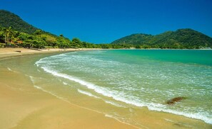 A beach with clear turquoise water, sandy shore, and lush green hills in the background. - Suíte Lázaro (Ubatuba)