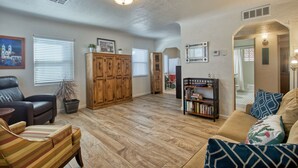 A spacious living room with hardwood flooring, a leather sofa, a striped armchair, a wooden cabinet, and a bookshelf. - Adorable 2 BR at the heart of town! (Las Cruces)