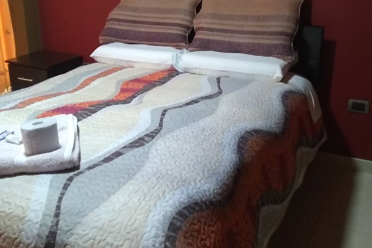 Una cama con un edredón estampado, dos almohadas y una toalla enrollada sobre una mesita de noche.
