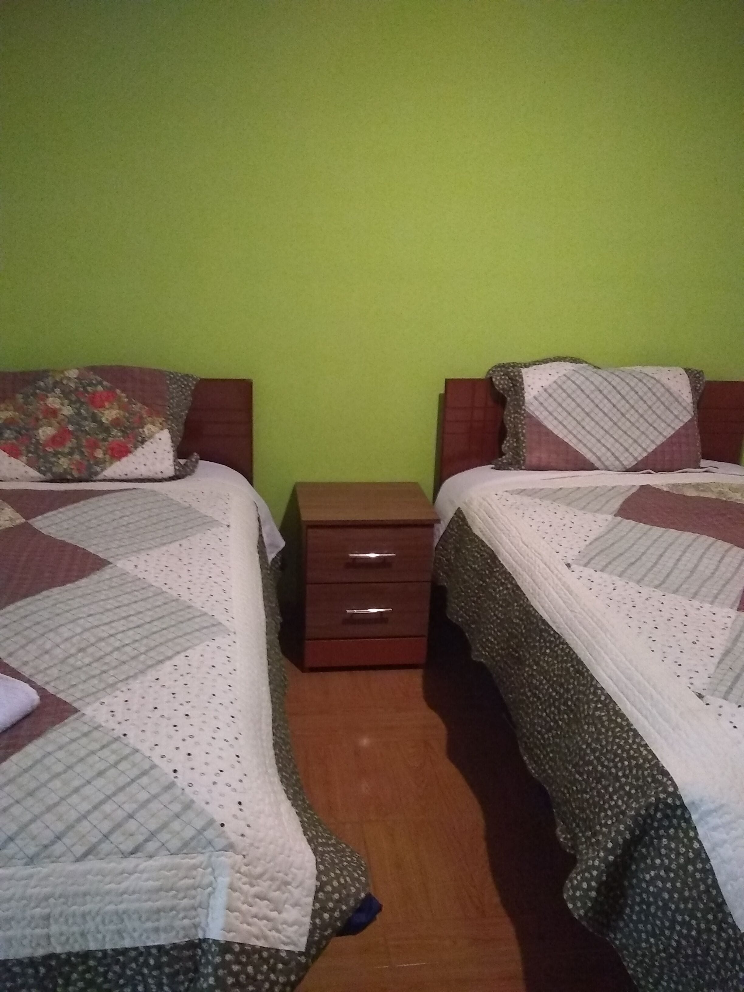 A room with two beds, each with a quilted bedspread and a pillow. There is a wooden nightstand with drawers between the beds.