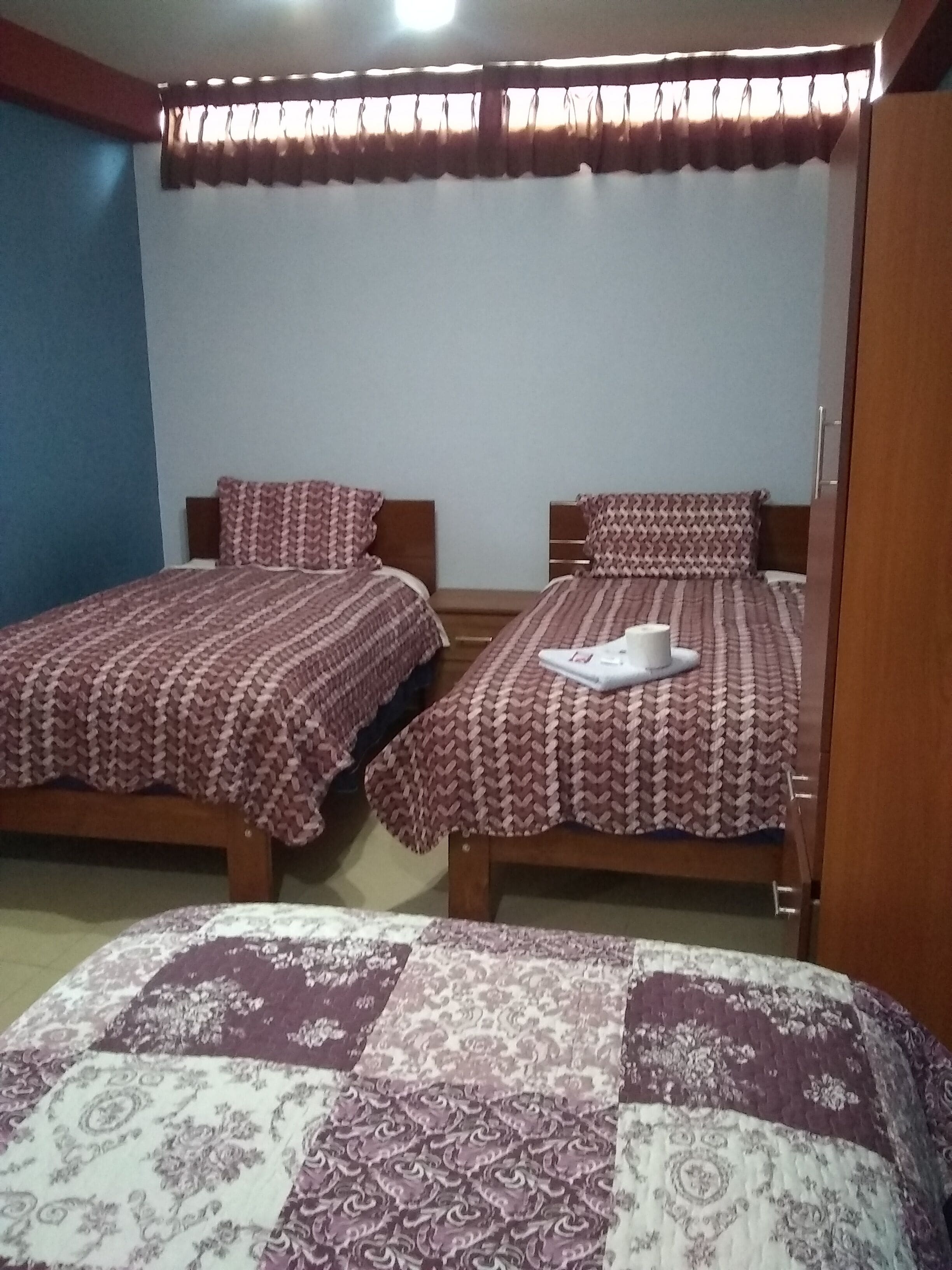 Quarto com duas camas de solteiro, cada uma com roupa de cama e almofadas estampadas, cabeceira de madeira e janela com cortinas.