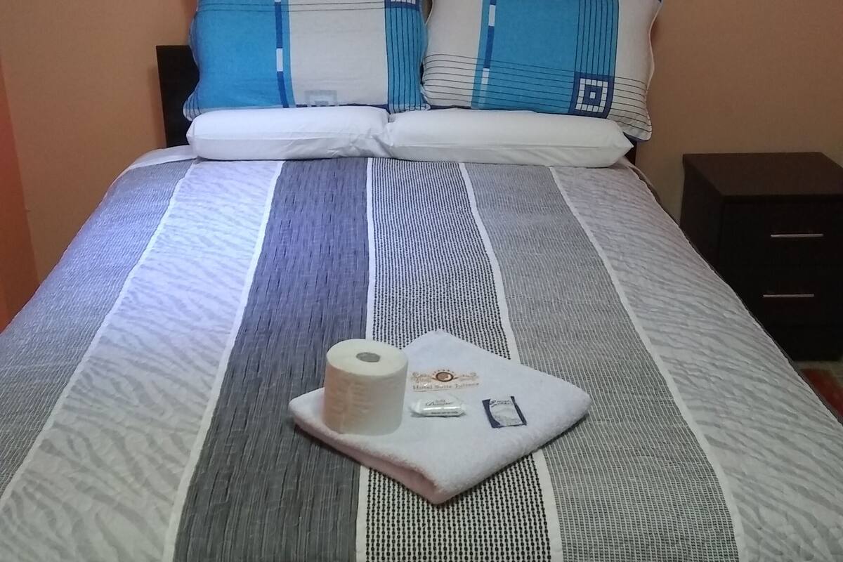 Una cama bien tendida con ropa de cama a rayas, una almohada con diseño geométrico y una mesita de noche con un cajón negro.