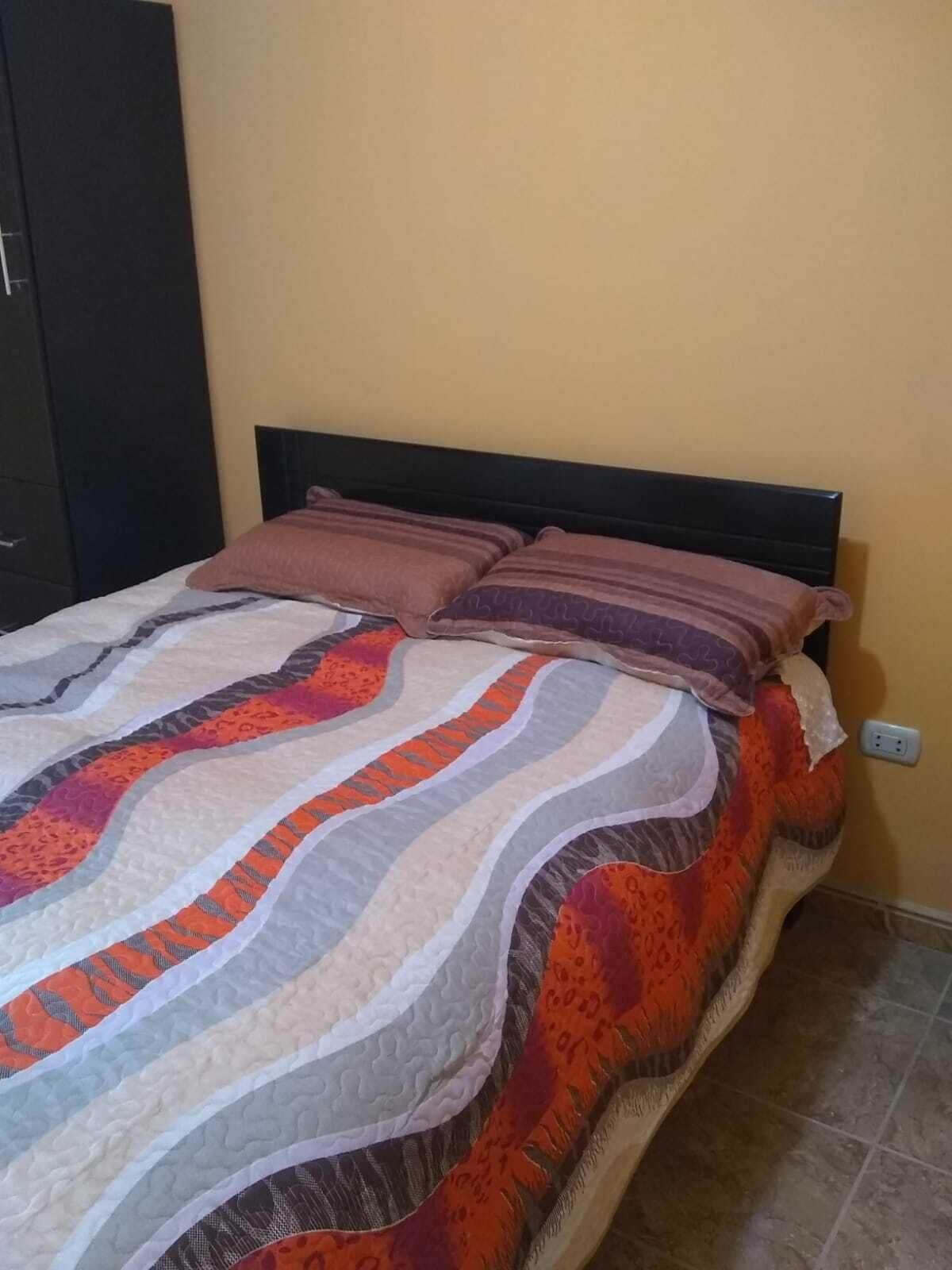 A bed with a patterned bedspread and two pillows, a dark headboard, and a wall socket.