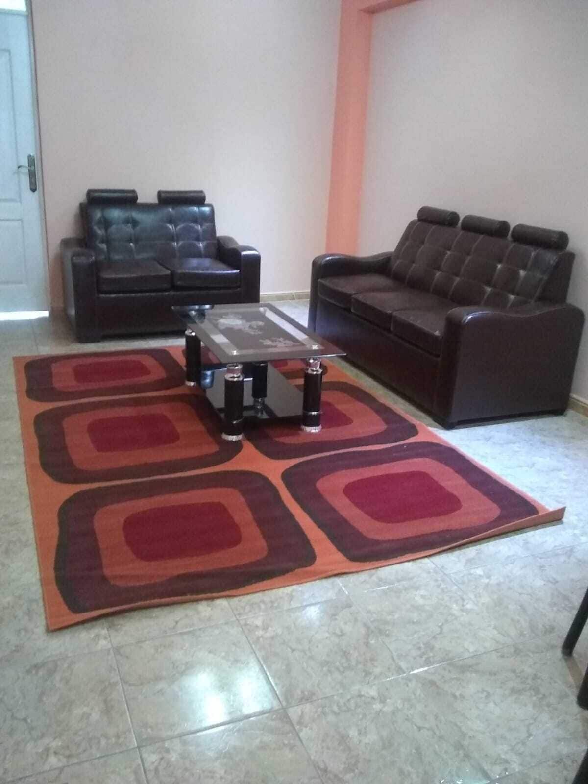 A living room with two dark leather sofas, a glass coffee table, and a patterned rug on a tiled floor.