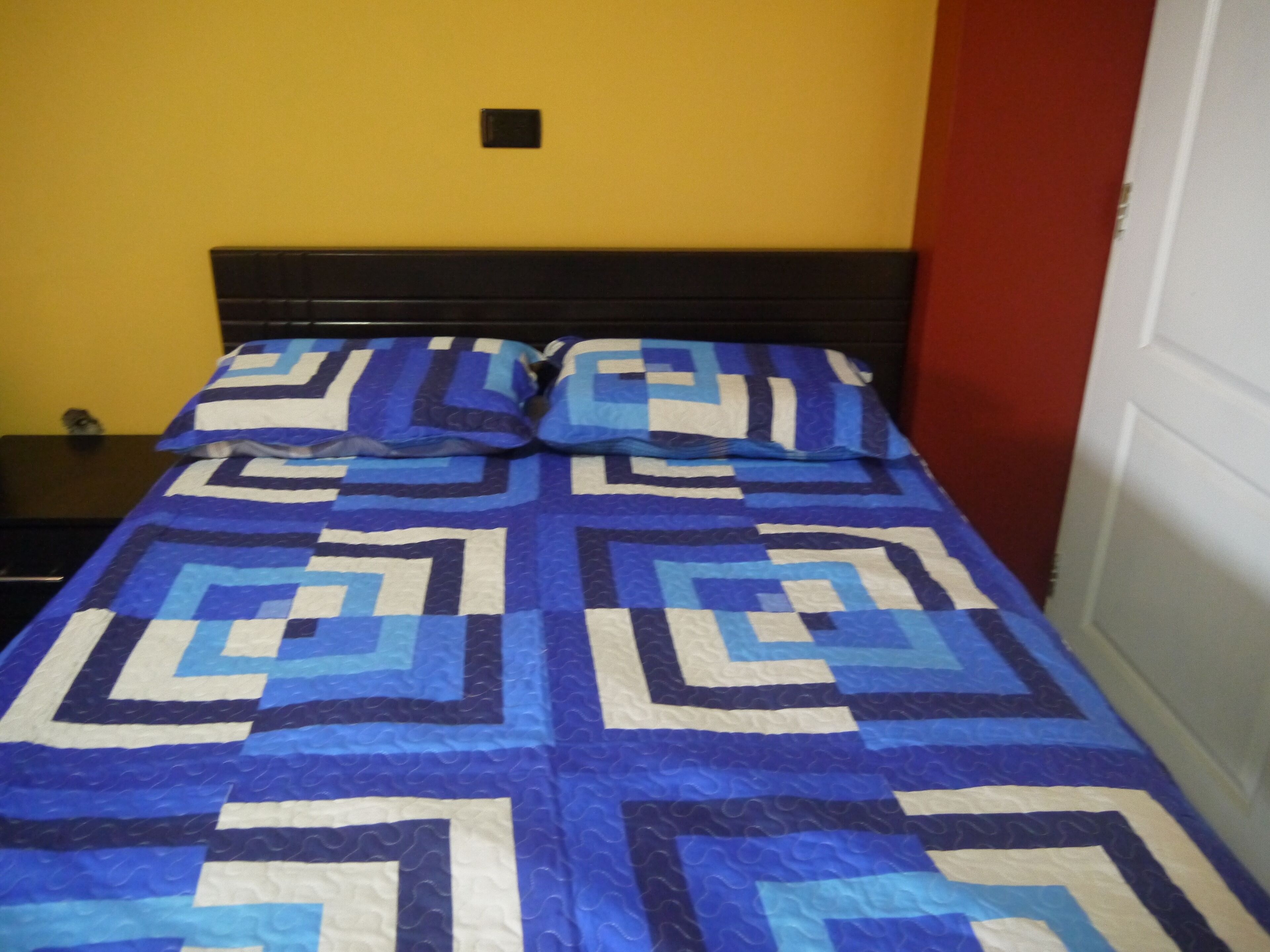 A bed with a geometric patterned bedspread, a black headboard, and a yellow wall with a red door.