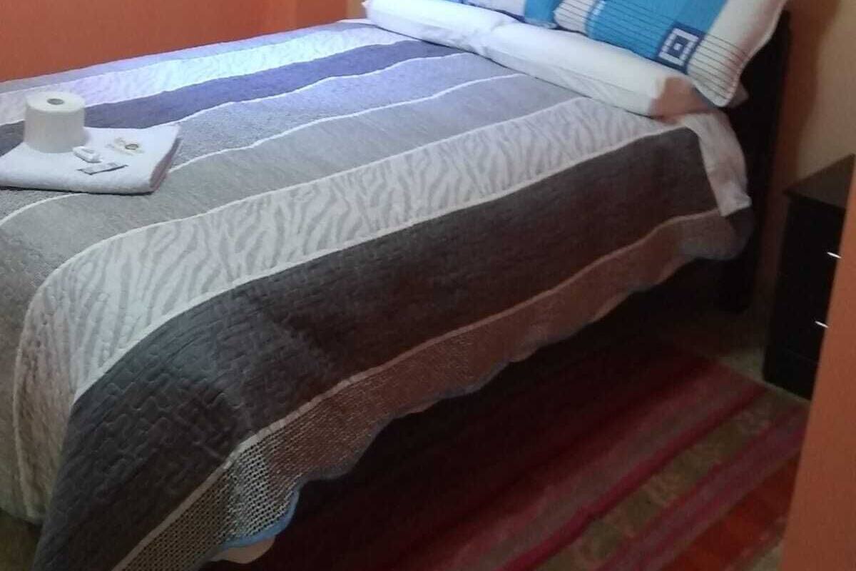 Una cama pequeña y bien hecha con una colcha a rayas y almohadas, una mesita con una caja de pañuelos, y una alfombra a rayas en el suelo.