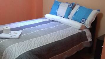 Una cama pequeña y bien hecha con una colcha a rayas y almohadas, una mesita con una caja de pañuelos, y una alfombra a rayas en el suelo.