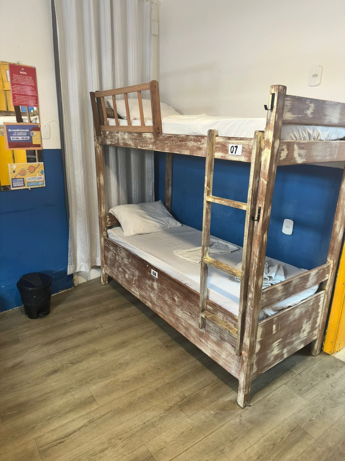 A bunk bed with wooden railings, a small ladder, and a white mattress. There is a black trash bin and a blue wall with a sign on it.