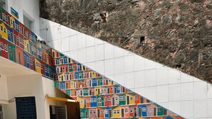 En patio med et rundt bord og fire gule stole, et farverigt muralmaleri af huse på væggen, og en potteplante.