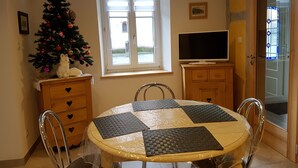 A dining area with a round table covered in plastic, four chairs, a Christmas tree with ornaments, a white dog figurine, a television on a wooden cabinet, and a window with blinds. - Charming spacious apartment for 4 people (Neuwiller les Saverne)