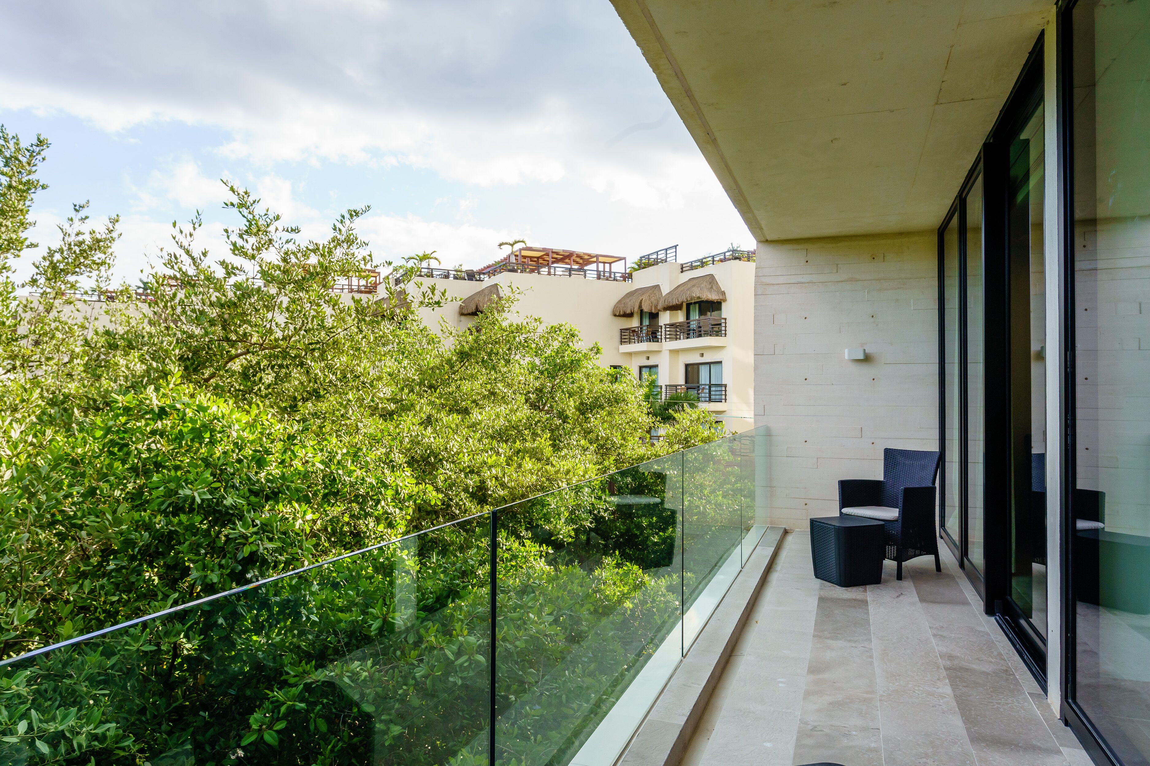 A balcony with glass railings overlooking a lush green area and a building with balconies.