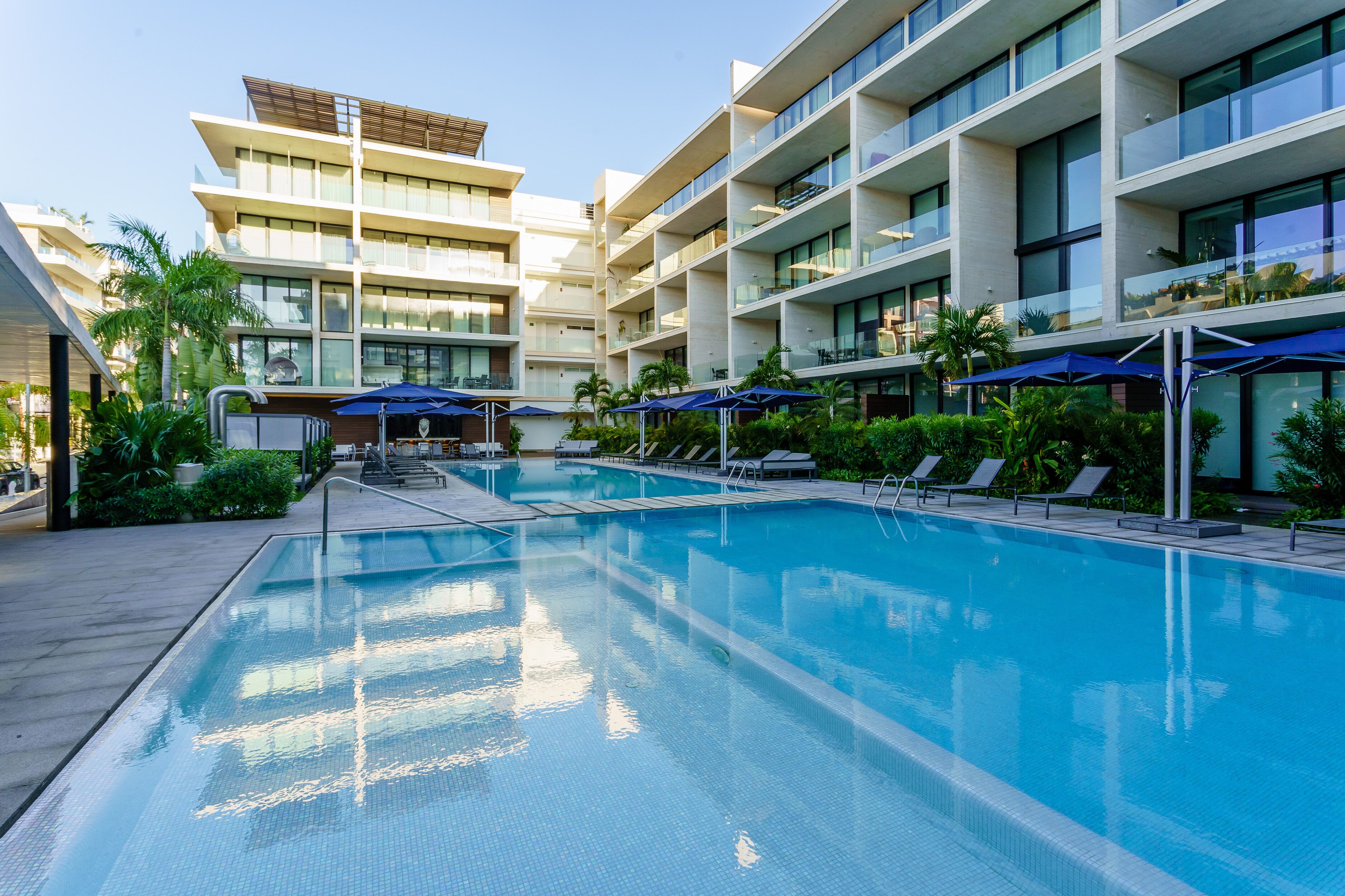 A swimming pool with lounge chairs and umbrellas, surrounded by modern buildings with balconies.