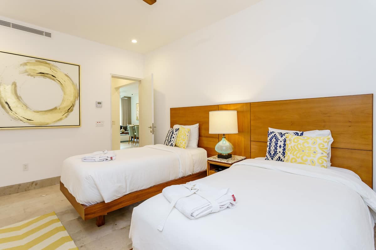 Quarto de hotel com duas camas, cabeceira de madeira, mesa de cabeceira com abajur e uma grande pintura abstrata na parede.