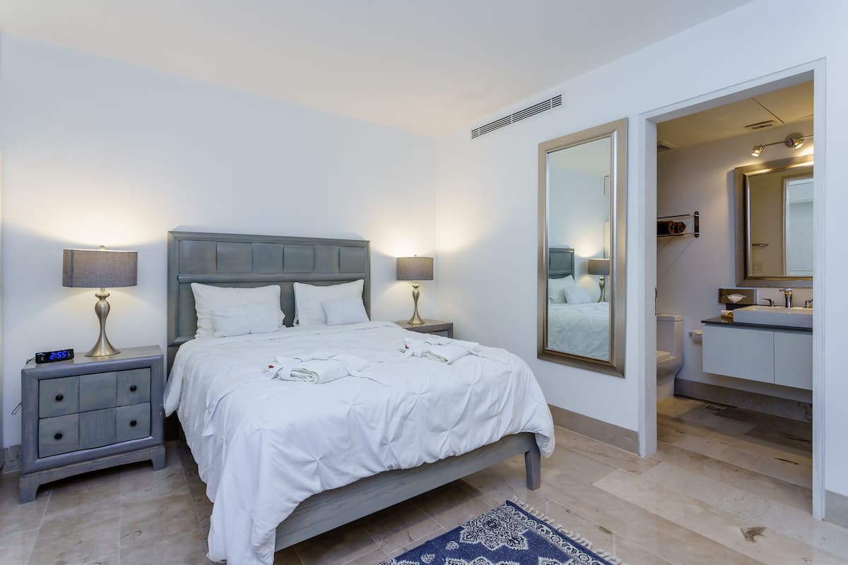 Quarto com cama, mesas de cabeceira, um espelho e um banheiro visível por uma porta aberta.