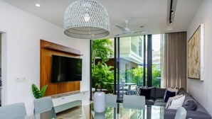 Een moderne woonkamer met een glazen eettafel, grijze banken, een grote televisie en een plafondventilator.