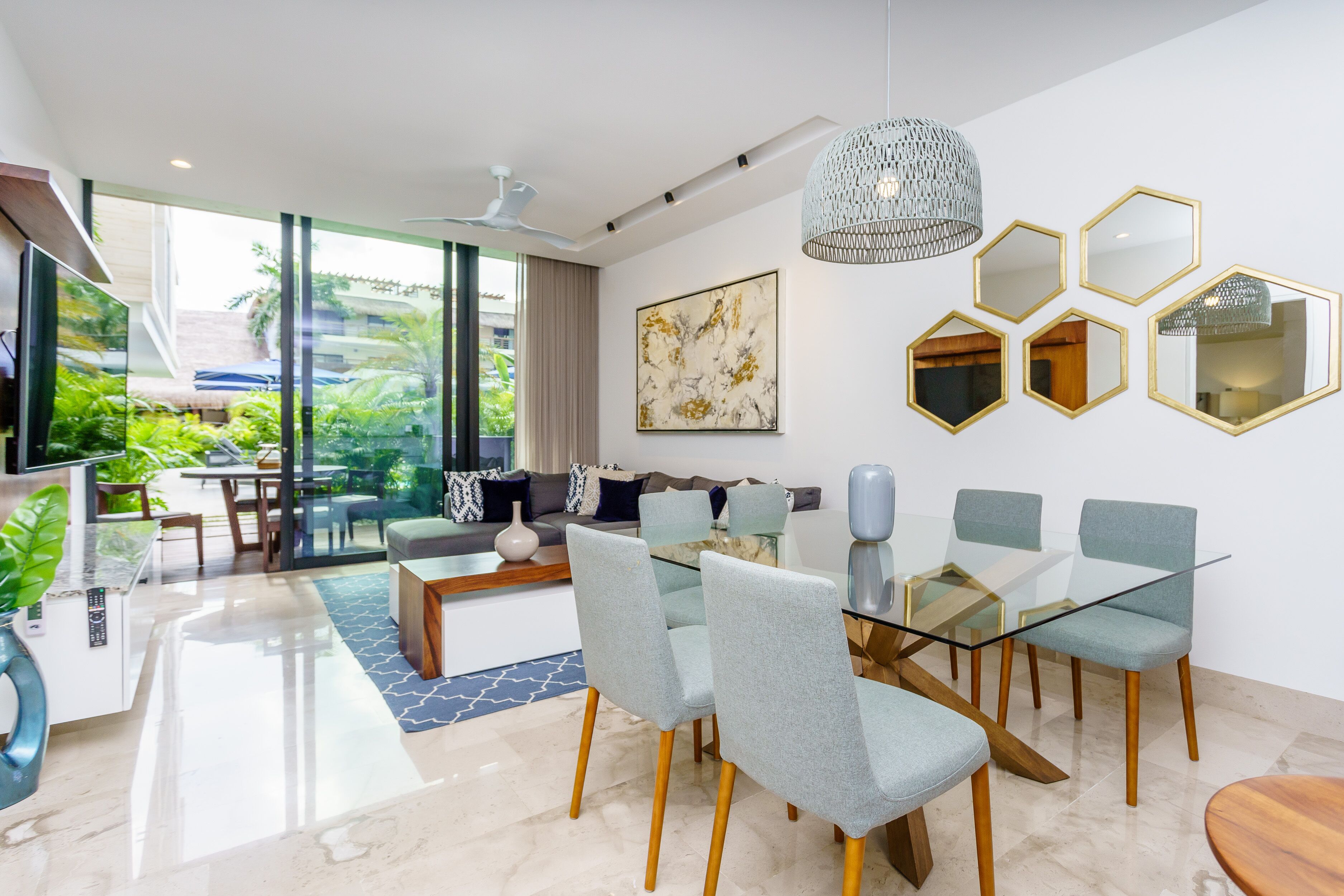 Una sala de estar moderna con una mesa de comedor de vidrio, sillas tapizadas en gris, un ventilador de techo y un ventanal que ofrece vistas a zonas verdes.