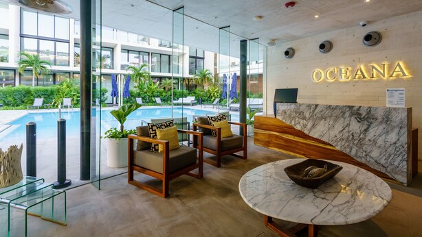 Een moderne hotel lobby met een receptiebalie, een zithoek en uitzicht op een zwembad.