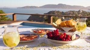 A table with plates of food, a glass of drink, and a view of the sea and hills in the background. - TOOL. BUZIOS. (Armaçao dos Buzios)