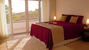 A hotel room with a bed, two bedside lamps, a white fluffy rug, and a view of greenery through a large window. - TOOL. BUZIOS. (Armaçao dos Buzios)
