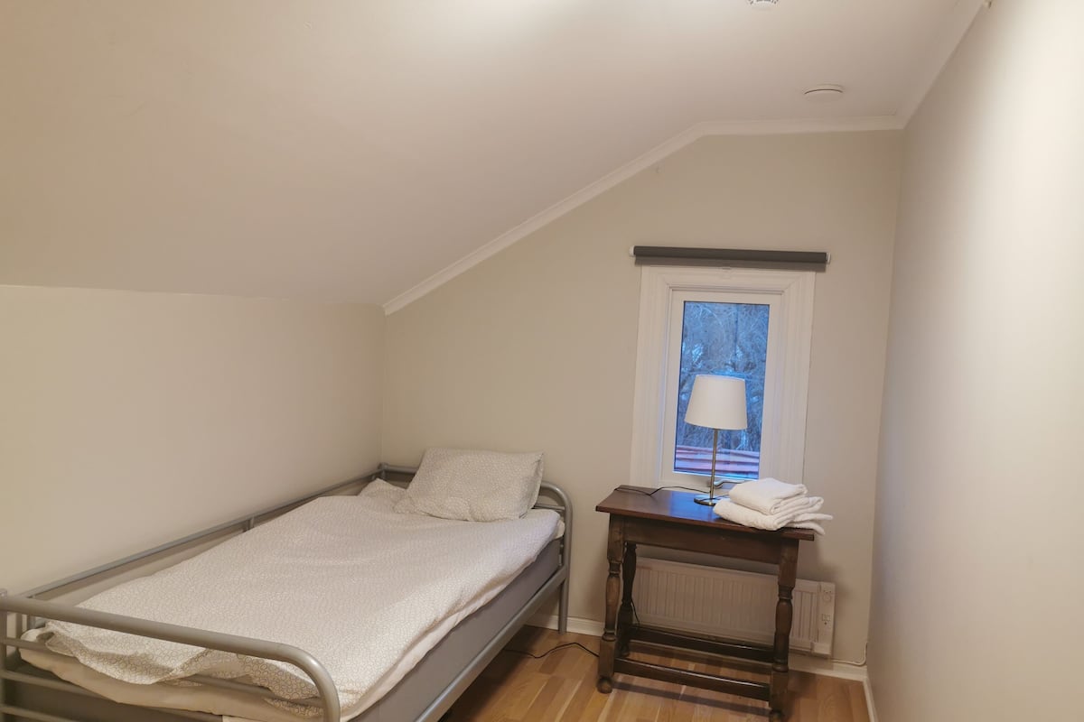 A small, single-bed room with a wooden floor, a window with a view of trees, and a bedside table with a lamp and towels.