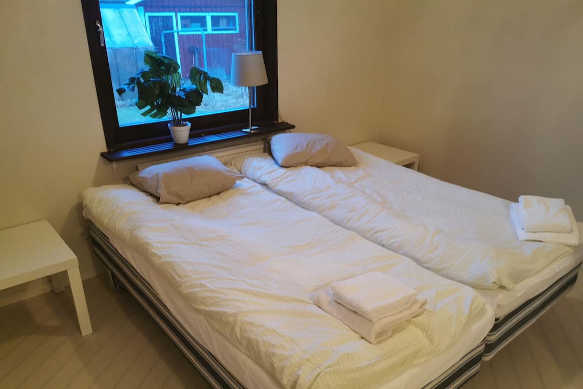 A double bed with white linens, a bedside table, a lamp, and a window with a view of a building and a plant.