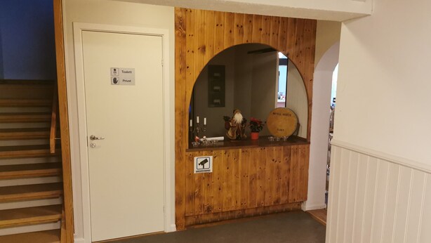 Interior de madera con una puerta blanca que dice 'Toilet' y 'Privat', un pequeño mostrador de recepción con el letrero 'MOTELL GASTIS FLOBY' y un espejo encima.
