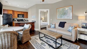 Cozinha moderna com geladeira, micro-ondas e cafeteira, sala de jantar com cadeiras de vime e mesa, sala de estar com sofá e mesa de centro, e ventilador de teto.