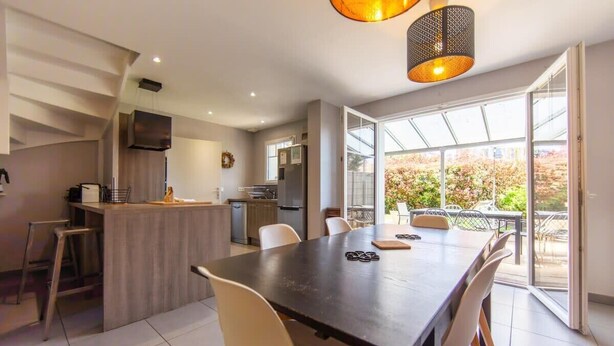 A modern kitchen with a central island, a dining area with a large table, and a view of a garden through a glass door.