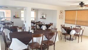 Uma sala de jantar com várias mesas e cadeiras, um ventilador de teto e uma janela com persianas.