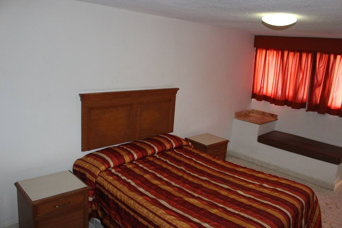 A bedroom with a wooden headboard, a striped bedspread, a bedside table, and a window with red curtains.