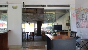 Una oficina con escritorio, sillas y una computadora. Hay una puerta de vidrio que da a un espacio exterior con mesas y sillas.