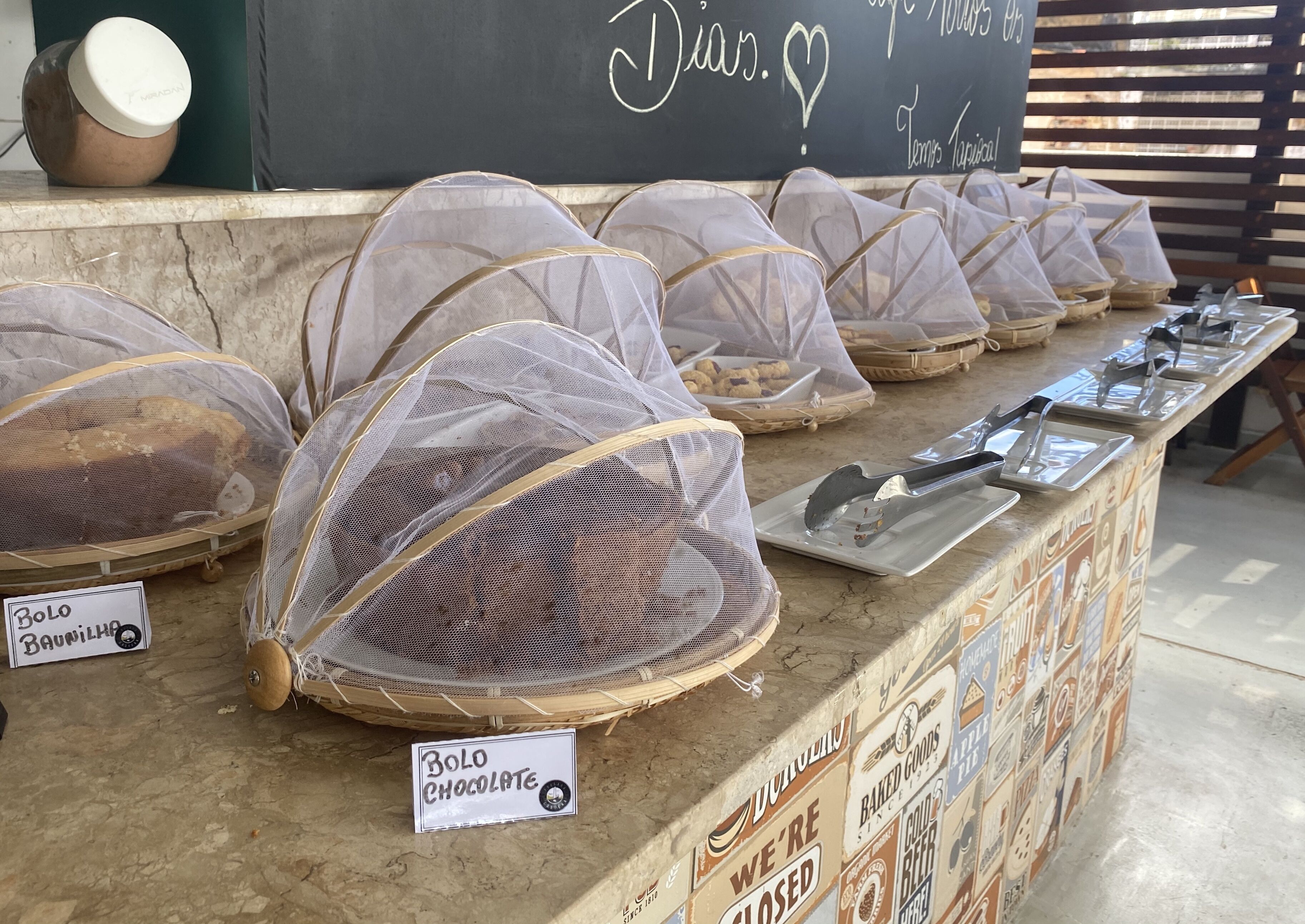 Exhibición de productos horneados cubiertos con etiquetas, una pizarra con mensajes y una pared de azulejos con varios letreros.