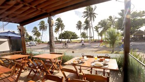 An outdoor seating area with wooden tables and chairs, overlooking a street with palm trees and a cyclist.