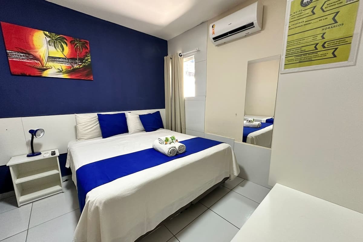 A hotel room with a bed, a nightstand, a desk lamp, a painting on the wall, an air conditioning unit, and a mirror.